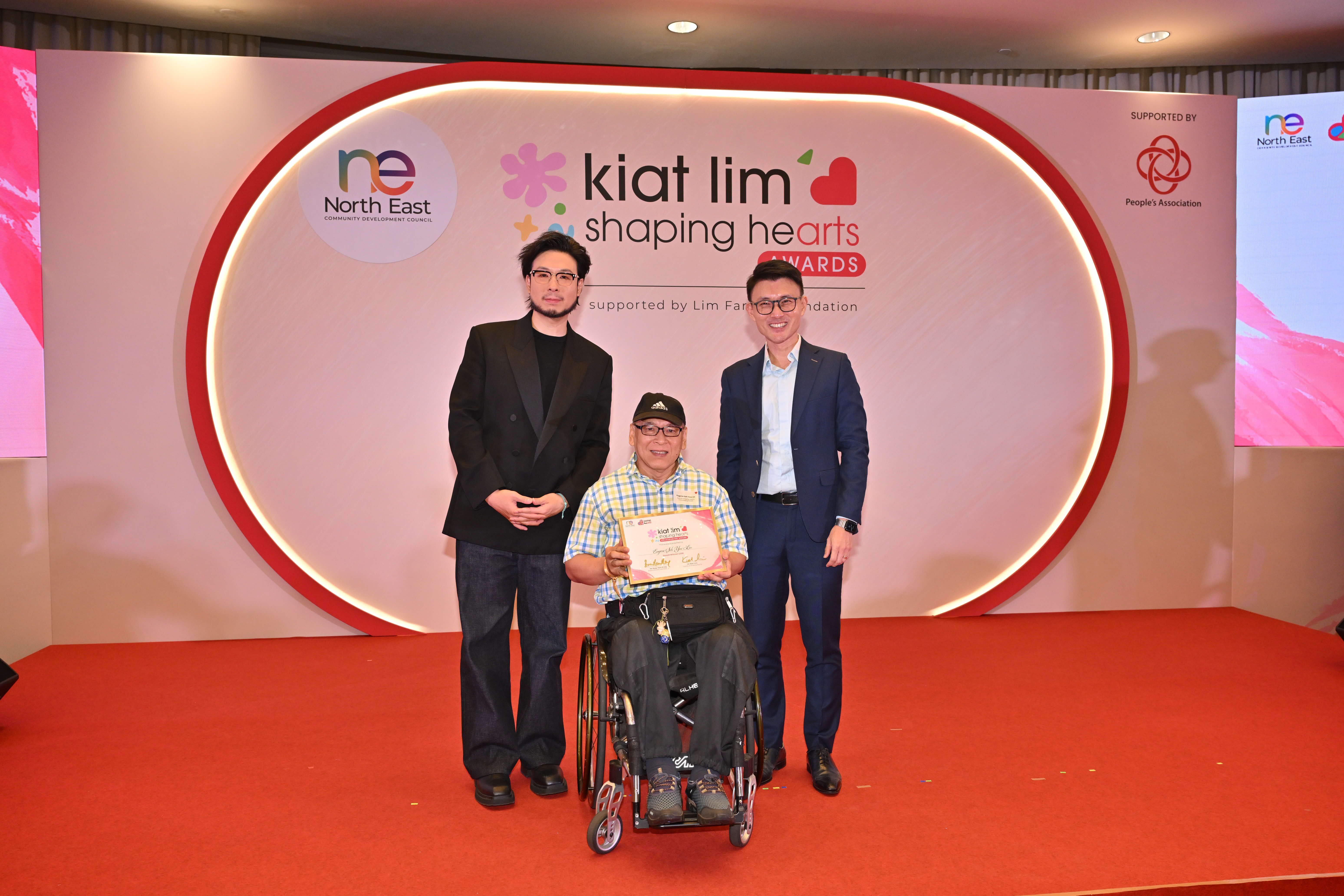 Award recipient standing on stage with the presenter, holding their certificate and posing for a group photo during the award ceremony, with a backdrop reading ‘Kiat Lim-Shaping Hearts Awards'.